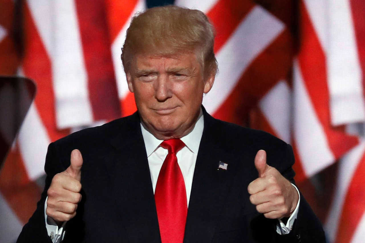 President Trump holding both thumbs up in front of US flags.